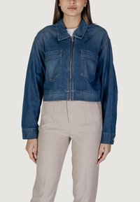 Giubbotto di jeans accorciato di colore blu scuro con chiusura a zip, due tasche sul petto e maniche lunghe. Indossato sopra pantaloni chiari.