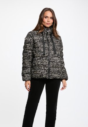 Femme portant une veste zippée texturée noire et blanche avec des cordons noirs et un pantalon noir sur un fond blanc uni.