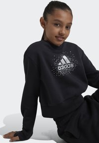 Sudadera negra con cuello redondo y mangas largas, que presenta un logo de Adidas blanco con un diseño moteado en la parte frontal. Tela suave.