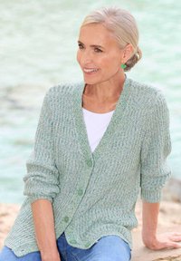 Hellgrün gestrickte Strickjacke mit V-Ausschnitt, gerippter Textur, drei Knöpfen und aufgerollten Ärmeln, getragen über einem weißen Oberteil und Jeans.