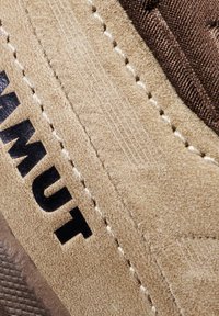 Close-up van een beige suède schoensectie met zwart "MUT"-lettering en witte stikseldetails op een textiel met structuur.