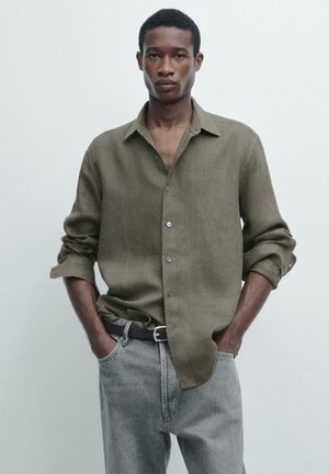 Mann trägt ein olivgrünes Hemd mit Knöpfen, die Ärmel sind hochgekrempelt, und graue Jeans, steht mit den Händen in den Taschen vor einem einfarbigen Hintergrund.