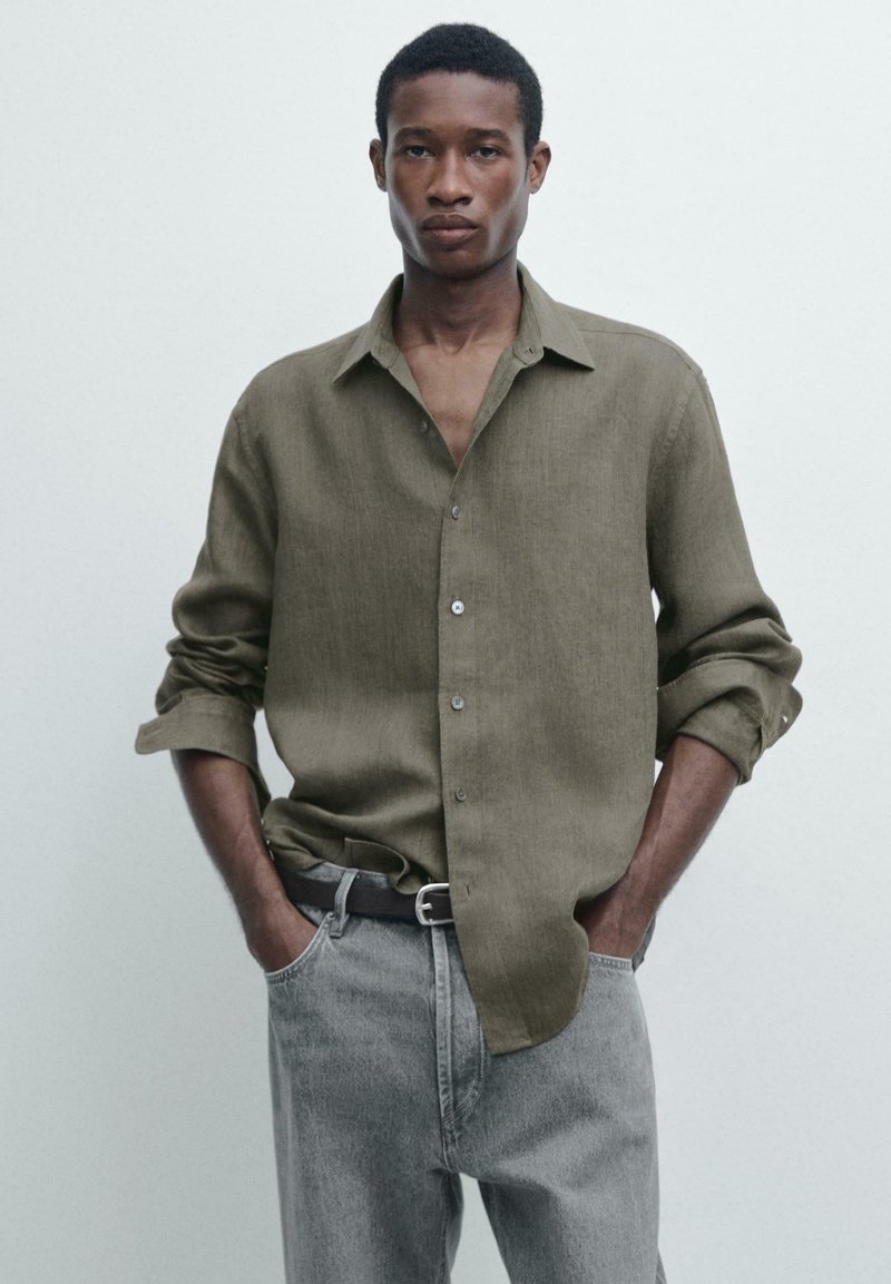 Mann trägt ein olivgrünes Hemd mit Knöpfen, die Ärmel sind hochgekrempelt, und graue Jeans, steht mit den Händen in den Taschen vor einem einfarbigen Hintergrund.