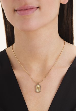 Woman wearing a gold chain necklace with a rectangular gold pendant framed by small gemstones, dressed in a black top.