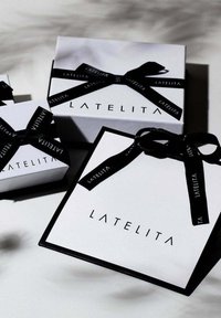 Tres cajas de regalo apiladas, con un exterior blanco y detalles en negro. Cada una tiene una cinta negra con la inscripción "LATELITA". Las cajas varían en tamaño.