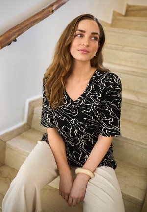 Kobieta o długich brązowych włosach siedząca na beżowych schodach, ubrana w czarną, wzorzystą bluzkę z krótkim rękawem, kremowe spodnie oraz kremową bransoletkę na lewej ręce.