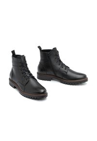 Botas chelsea de cuero negro con un acabado texturizado, puntera redonda, cierre de cordones, cremallera lateral y suela resistente. Detalles en color marrón en el borde de la suela.