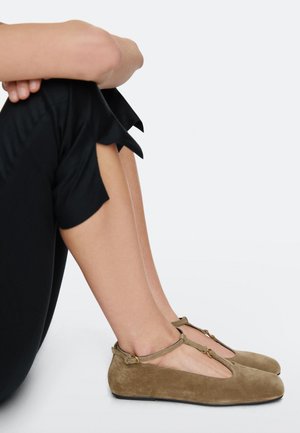 Kvinde siddende med bøjede ben, iført sorte cropped bukser med bølgekantede kanter og beige flade T-remme sko i ruskind.