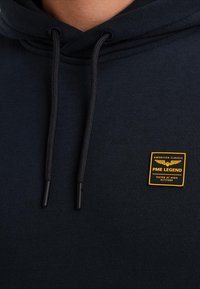 Detailní záběr na černou mikinu s kapucí, černými šňůrkami a žlutým čtvercovým štítkem s nápisem "PME Legend Testováno ve velkých výškách."