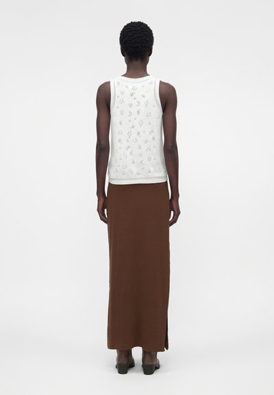 Άτομο που φοράει λευκό αμάνικο τοπ με μικρά μαύρα ουράνια και αφηρημένα σύμβολα, συνδυασμένο με μακριά καφέ φούστα και μαύρα παπούτσια.