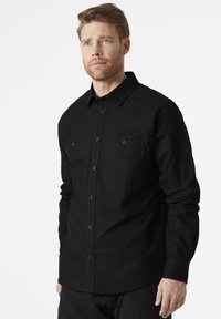 Zwarte overhemddblouse met lange mouwen, een enkele borstzak en bijpassende knopen. Gemaakt van een stevige, gladde stof met minimale stikdetails.