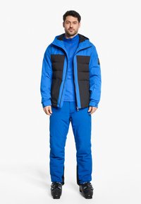 Blaue gepolsterte Jacke mit schwarzen Akzenten, kombiniert mit einem blauen Basisausrüstungsstück und passenden blauen Hosen. Texturiertes Material und funktionelles Design.