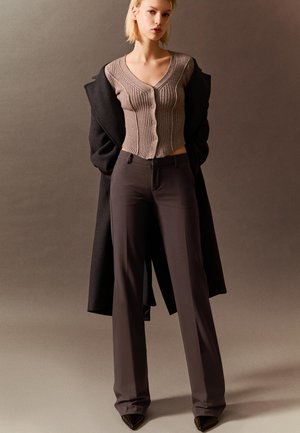 Beige ribbstickad kofta med knappar, kombinerad med mörkbruna skräddarsydda byxor och en svart lång kappa. Spetsiga svarta klackar förstärker looken.
