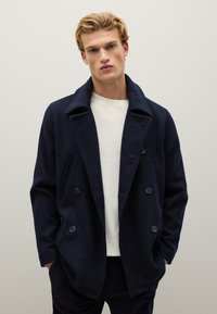 Cappotto peacoat in lana blu navy con chiusura doppiopetto, ampi revers, tasche laterali e tessuto testurizzato. Indossato sopra un maglione crema chiaro.