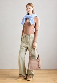 Femme portant un pull en V marron avec une écharpe bleue, un pantalon large beige, tenant un sac à main en daim taupe, se tenant sur un sol en bois.