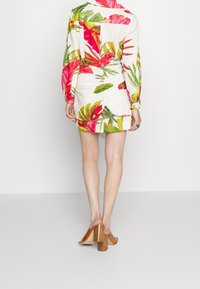Blumenkleid mit langen Ärmeln, das lebendige rosa und grüne Muster auf einem hellen Hintergrund zeigt. Getragen mit beige Blockabsatzschuhen.
