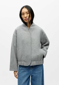 Veste en mélange de laine grise avec un col montant, des épaules tombantes, des poches latérales et un ourlet ajusté. Associée à un jean en denim bleu.