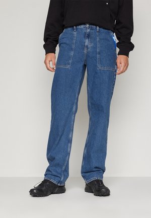 Man met blauwe spijkerbroek met grote voorzakken, zwarte sneakers en een zwart shirt met lange mouwen tegen een effen achtergrond.