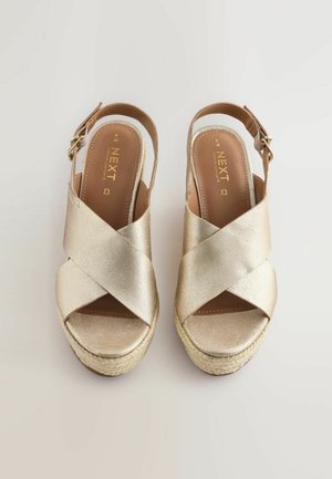 Zelta sintētiskie sandales ar atvērtiem papēžiem un krustenisku jostu dizainu, espadrillu tipa jutu zoli un regulējamu sprādzi. Gluda tekstūra un mīkstinātas iekšējās zoles.