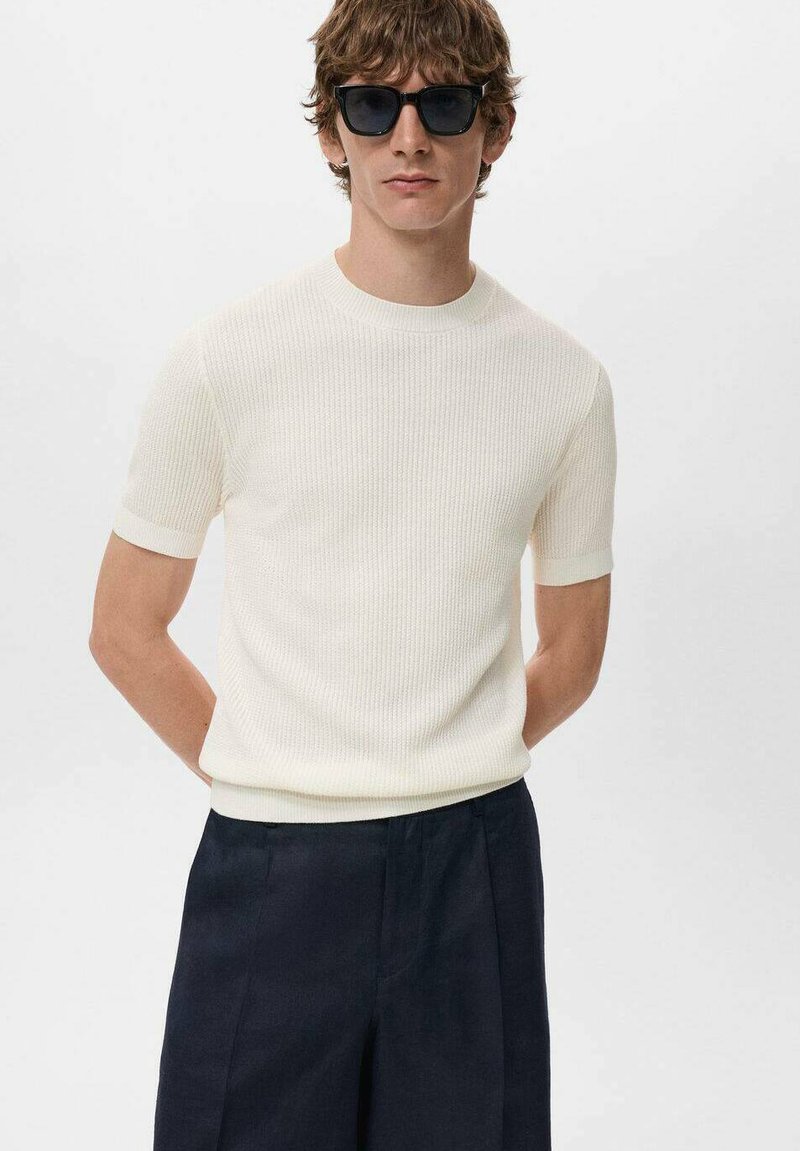 Suéter de punto acanalado crema de manga corta con cuello redondo, combinado con pantalones azul marino y gafas de sol negras. Textura suave, diseño ajustado.