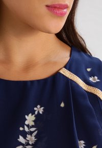 Marinblus med blommönster, guldaccent längs halsringningen och lätt material. Närbild av axelområdet.