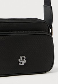 Borsa rettangolare nera in tessuto strutturato, con logo argentato, chiusura con cerniera e tracolla regolabile.