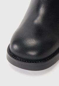 Bottine en cuir noir avec un bout arrondi. Présente une finition texturée et une semelle en caoutchouc solide et plate. Couture décorative le long du bord.