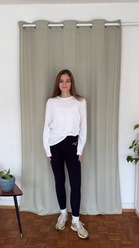 Fehér hosszú ujjú ing, fekete leggings logóval, és szürke sportcipő. Egy világoszöld függöny előtt áll, mellette egy cserepes növény.