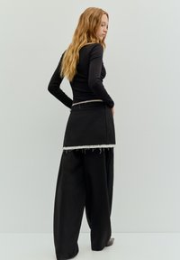 Sort langærmet top kombineret med en sort nederdel med høj talje og frynset kant, suppleret med løstsiddende sorte bukser.