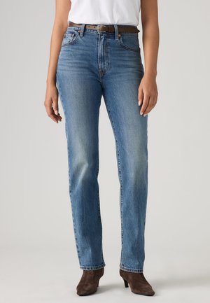Vrouw die lichtblauwe jeans met rechte pijpen, wit t-shirt, bruine riem en bruine enkellaarsjes met hak draagt, staand tegen een effen achtergrond.