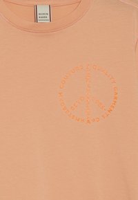 Pfirsichfarbiges Baumwoll-T-Shirt mit einem runden, orangefarbenen Grafiktextdesign mit Markennamen und Gründungsdetails. Klassischer Rundhalsausschnitt.