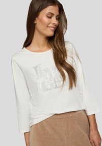 Frau mit langen braunen Haaren, die ein weißes Langarmshirt mit dem Text "IMAGE" und beige Cordhosen trägt, lächelt und schaut nach unten.