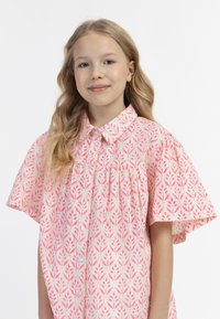 Camicia corta rosa chiaro con maniche corte, caratterizzata da un motivo floreale in rosa scuro, colletto con bottoni e dettagli arricciati al busto.