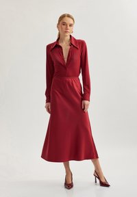 Largo vestido de satén rojo con cuello, botones en la parte delantera y mangas largas. La falda se abre suavemente hasta el suelo. Lleva unos zapatos de tacón rojos a juego.