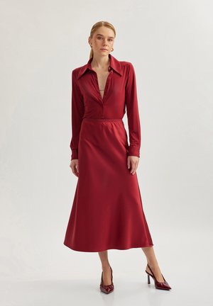 Lungo vestito rosso in satin con collare, chiusura a bottoni e maniche lunghe. La gonna si allarga delicatamente fino al pavimento. Indossato con tacchi abbinati rossi.
