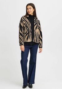 Veste en fausse fourrure beige avec des rayures noires zèbre ; présente une fermeture éclair, un col montant et une coupe décontractée. Portée sur un haut noir et un jean.