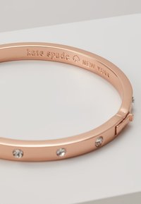 Rose gold bangle bracelet with a smooth finish, featuring three clear crystal accents and engraved brand name, hinged closure.