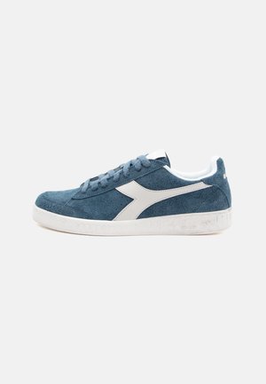 Blue suede sneaker with white accents, textured surface, round toe, and rubber outsole. Features laces and a logo on the side.