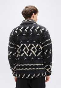 Μαύρο και κρεμ φλις μπουφάν με γιακά, με υφή απαλή και γεωμετρικά σχέδια στο πίσω μέρος.