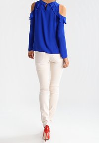 Blå kall-axlad blus med volangdetaljer; tillsammans med figurnära vita jeans och röda högklackade skor, sedd bakifrån.