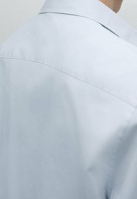 Camicia azzurro chiaro con una texture liscia, colletto a punta e sottili dettagli di cucitura, caratterizzata da un design aderente sul retro.