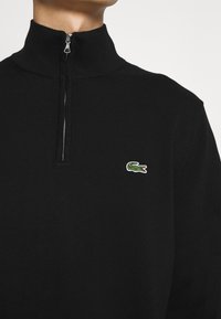 Svart stickad zip-up tröja med hög krage och en gr ön krokodil-logotyp på bröstet. Har en metallisk dragkedja och ribbad textur.