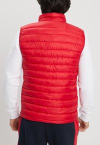 Gilet imbottito rosso trapuntato con colletto alto, texture liscia e cuciture orizzontali. Indossato sopra una camicia bianca a maniche lunghe.