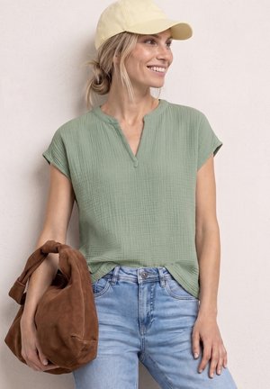 Femme portant une casquette jaune, un haut à manches courtes vert clair texturé, un jean bleu clair, tenant un sac en daim marron, souriante et regardant à droite.