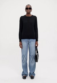 Maglione nero a maniche lunghe, jeans azzurri a vita alta con motivo logo e borsa nera. Modelli che indossano occhiali da sole scuri e ballerine.