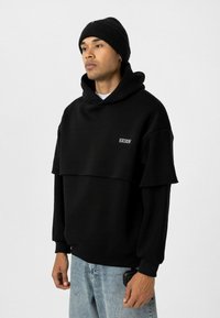 Un hoodie noir surdimensionné avec un design superposé, présentant un ourlet côtelé, des broderies à l'avant, associé à un jean bleu clair et un bonnet noir.