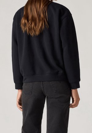 Frau mit langen braunen Haaren, die einen schwarzen Pullover und ausgewaschene schwarze Jeans trägt, von hinten vor einem schlichten Hintergrund gezeigt.