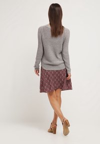 Šedý texturovaný svetr v kombinaci s vínově červenou vzorovanou sukní. Modelka má na sobě nahé podpatky. Souprava se vyznačuje uvolněným střihem a splývavým designem.