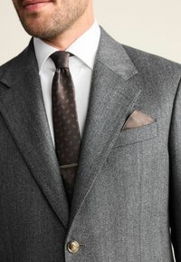 Grauer Fischgrät-Blazer mit einem Knopf, weißes Hemd und braune Krawatte mit blauen Mustern. Braunes Einstecktuch.