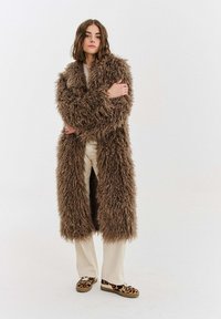 Hnědý dlouhý kabát z umělé kožešiny s texturovaným chlupatým materiálem, doplněný krémovými kalhotami a nazouvacími botami s leopardím potiskem.